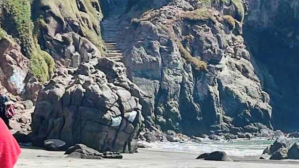 Un edificio costiero con facciata in pietra e tetto piatto, vicino a una scogliera rocciosa e una spiaggia.