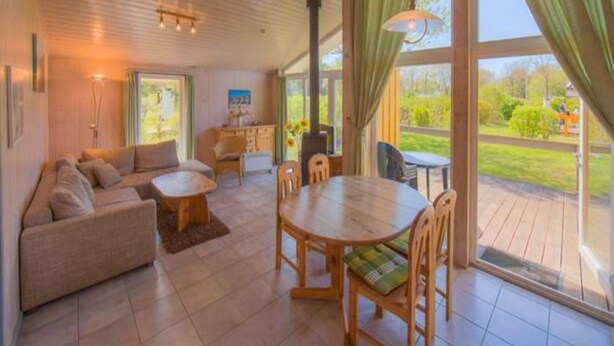 A spacious living room with a sofa, a dining table, and chairs. There is a large window with curtains, and a view of the outdoors. - Ferienhaus mit Sauna im Ferienpark Mirow (Mirow)