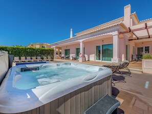 A hot tub with a wooden exterior, a pool, lounge chairs, and a building with a tiled roof.