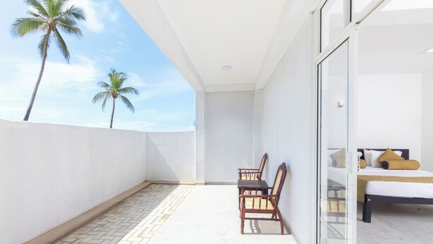 A balcony with a tiled floor, two wooden chairs, a bed with white bedding and a yellow pillow, and a view of palm trees against a clear sky. - Palazzo Beach Hikkaduwa (Hikkaduwa)