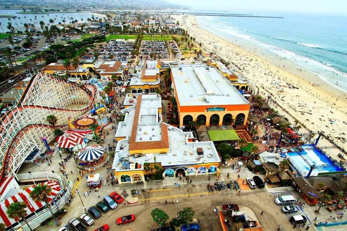 An aerial view of a coastal area with a beach, amusement park, and a large building with arches.