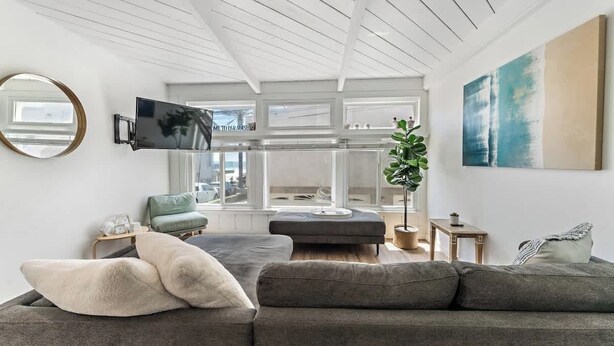 A modern living room with a grey sofa, a round mirror, a flat-screen TV, and a large window offering a view of the city. - Mission Beach w/ OceanViews Surfboards Parking AC (San Diego)