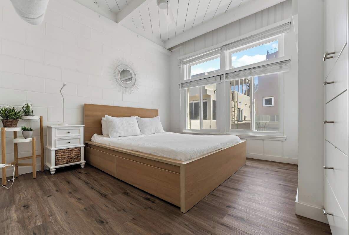 Una camera da letto con un letto di legno, pareti bianche e ampie finestre.
