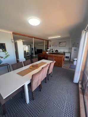 A dining area with a long table, chairs, a kitchen in the background, and a patterned carpet. - Beachfront Bliss with Stunning Eyre Peninsula Views (Miranda)