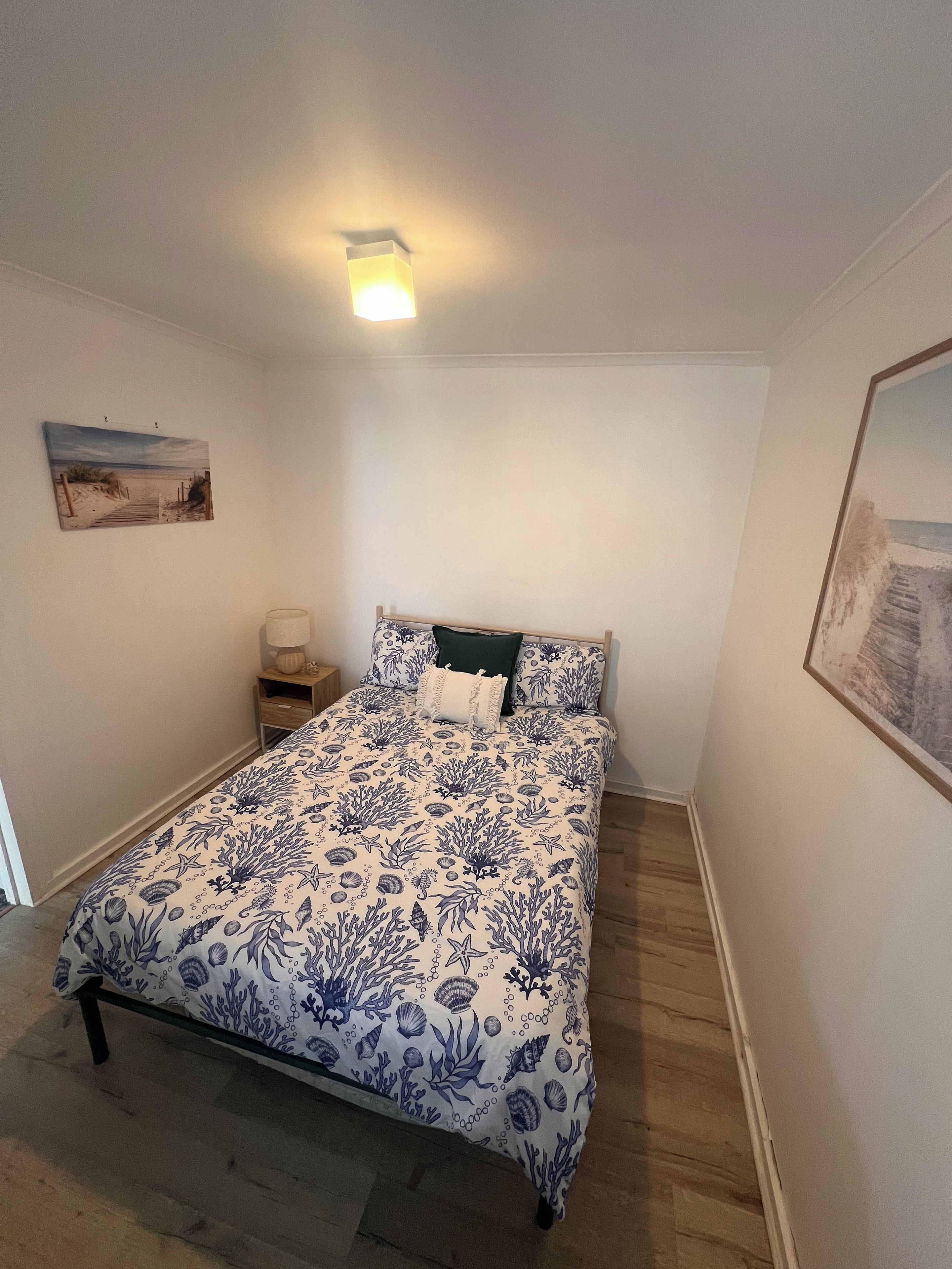 Een slaapkamer met een bed, nachtkastje, lamp en schilderij aan de muur.