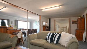 Ein Wohnzimmer mit Sofa, Sessel, Esstisch und Stühlen. Ein Kamin, ein Spiegel und ein Surfbrett sind ebenfalls vorhanden.