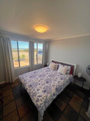 Un dormitorio con cama, ventana con cortinas, mesita de noche con ventilador y vistas al exterior.