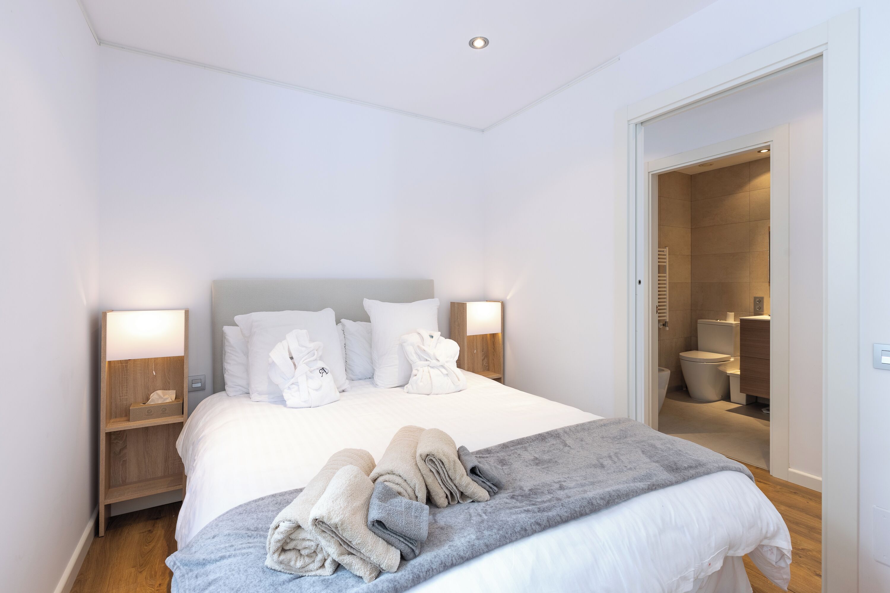 A hotel room with a neatly made bed, two bedside tables with lamps, a bathroom visible through an open door, and a grey throw blanket on the bed.