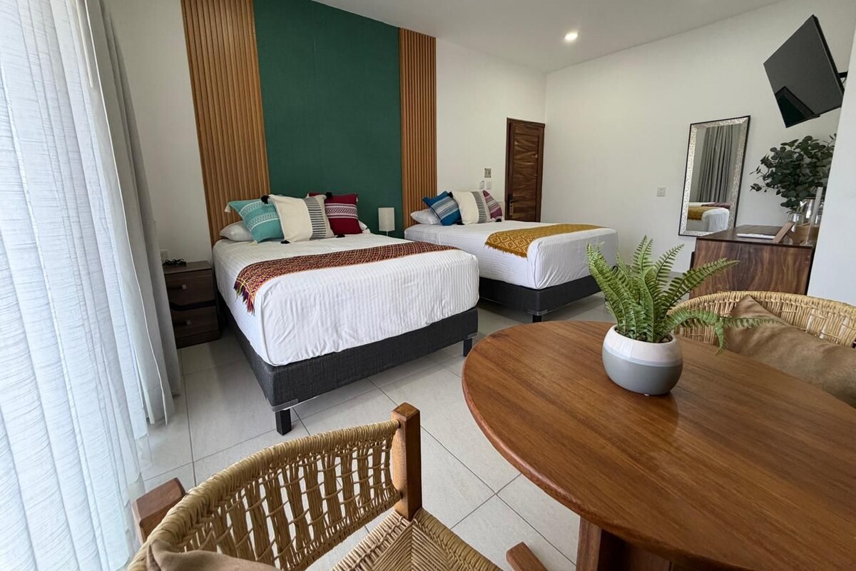 A hotel room with two beds, a dining table, a ceiling fan, and a potted plant.