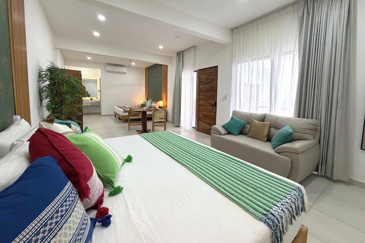 A hotel room with a bed, a sofa, a dining area, and a large window with curtains.