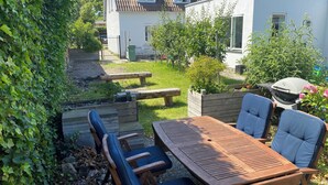Eine Terrasse mit Holztisch und -stühlen, einem Grill und einem kleinen Garten mit Bank und Holzzaun.