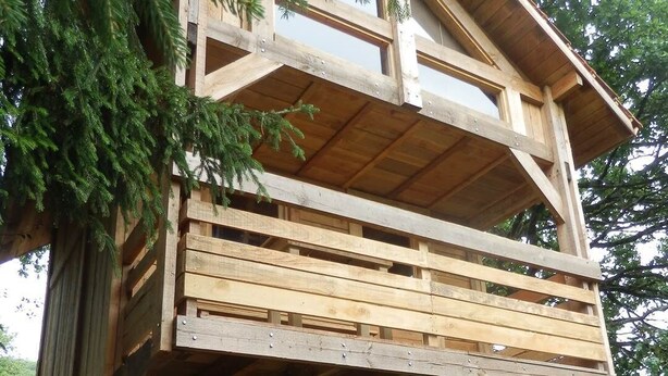 A wooden cabin with a balcony and a small window, surrounded by greenery.