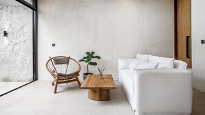 A modern living room with a white sofa, a wooden coffee table, a wicker chair, and a large window. - Blue Kai Villa by Alfred in Bali (Pecatu)