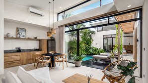 A modern kitchen with wooden cabinets, a dining area with a table and chairs, and a large sliding glass door leading to a pool area with plants. - Blue Kai Villa by Alfred in Bali (Pecatu)