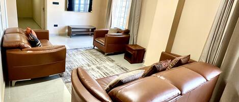 A modern living room with brown leather sofas, a flat-screen TV mounted on the wall, and a large window with curtains.