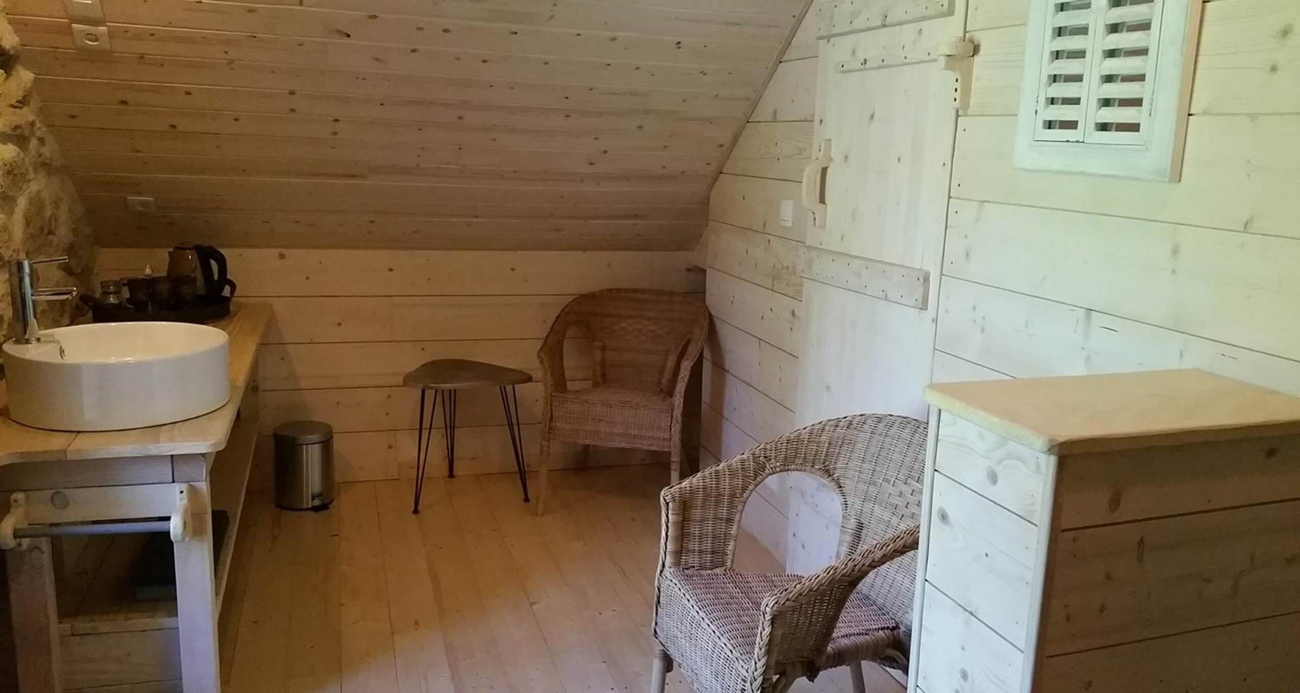 Una habitación pequeña y acogedora con detalles de madera, un lavamanos blanco, una silla de mimbre y una mesita.