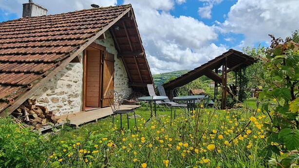 A rustic stone building with a sloped roof, a wooden door, and a small porch with outdoor seating. - Nature-themed Bed and Breakfast (Broye)