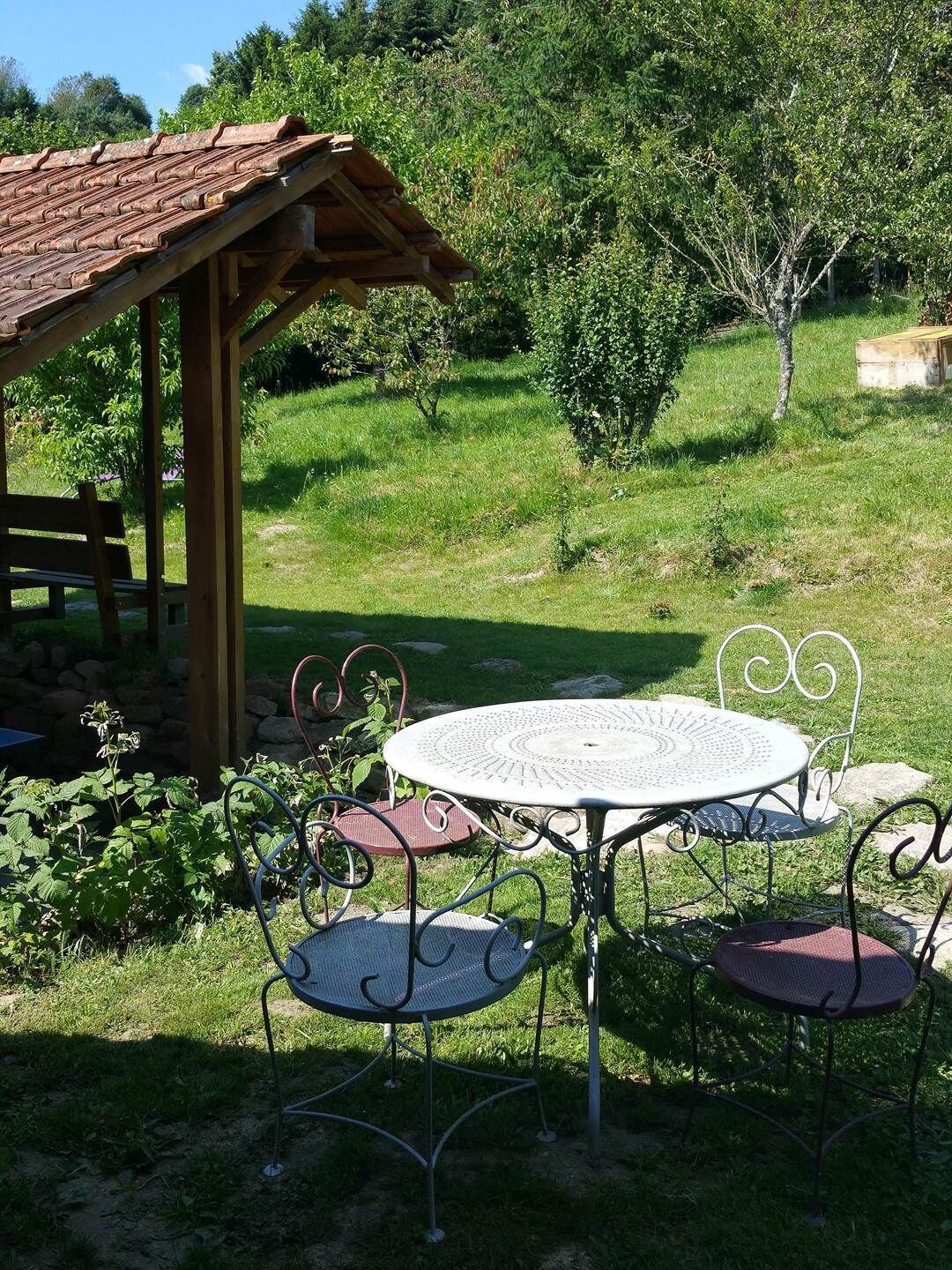 A wooden gazebo with a tiled roof, a round metal table with four chairs, and a grassy outdoor area with trees and shrubs.