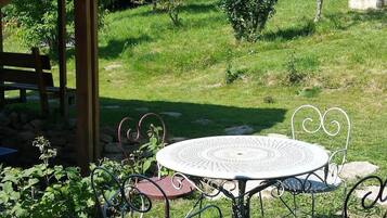 A wooden gazebo with a tiled roof, a round metal table with four chairs, and a grassy outdoor area with trees and shrubs.