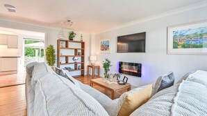 Ein modernes Wohnzimmer mit einer großen Couch, einem Holztisch und einem Flachbildfernseher. Ein Bücherregal mit Dekorationselementen und ein Gemälde sind an der Wand zu sehen.