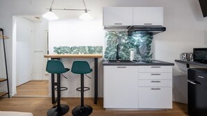 Una cocina compacta con armarios blancos, encimera negra y estufa moderna. Hay dos taburetes verdes y una lámpara colgante.