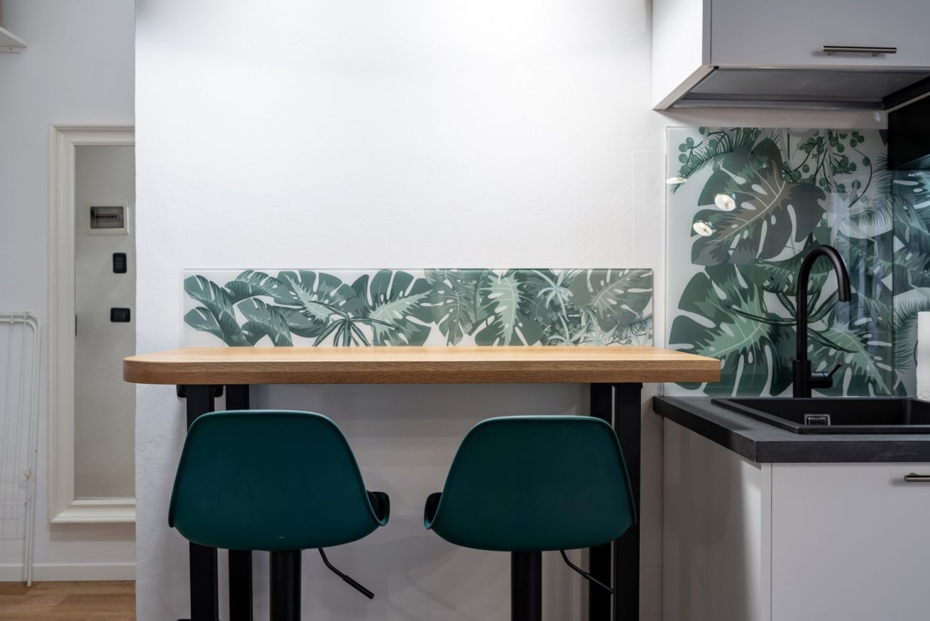 A kitchen with a wooden table, two green chairs, and a backsplash with tropical leaf patterns.