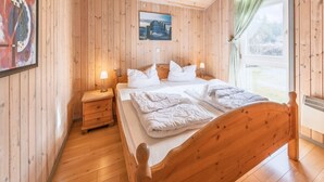 A wooden-paneled bedroom with a bed, bedside tables, a painting, and a window with curtains. - Ferienhaus mit Sauna im Ferienpark Mirow (Mirow)