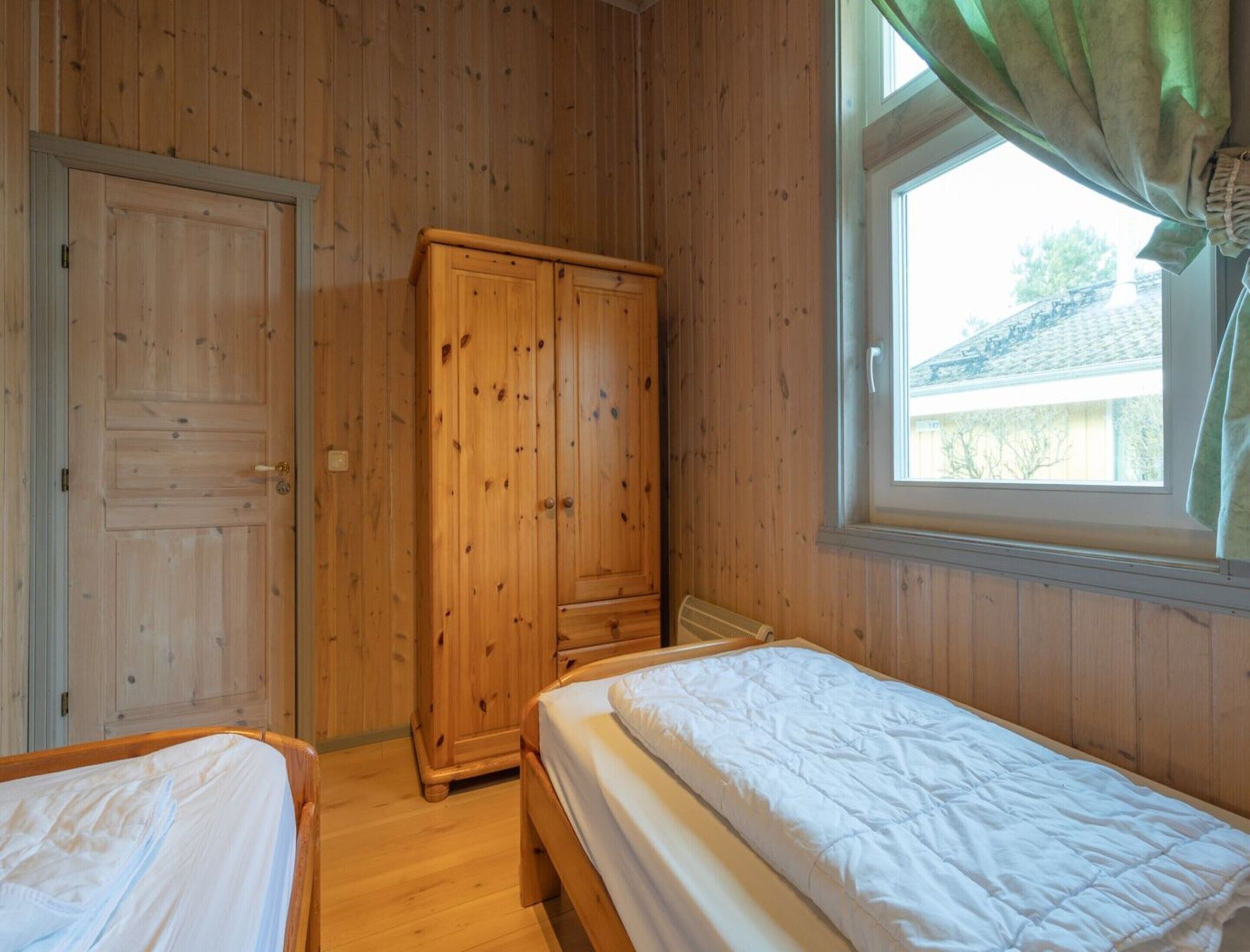 A room with wooden walls and floors, two beds, a wardrobe, and a window with curtains.