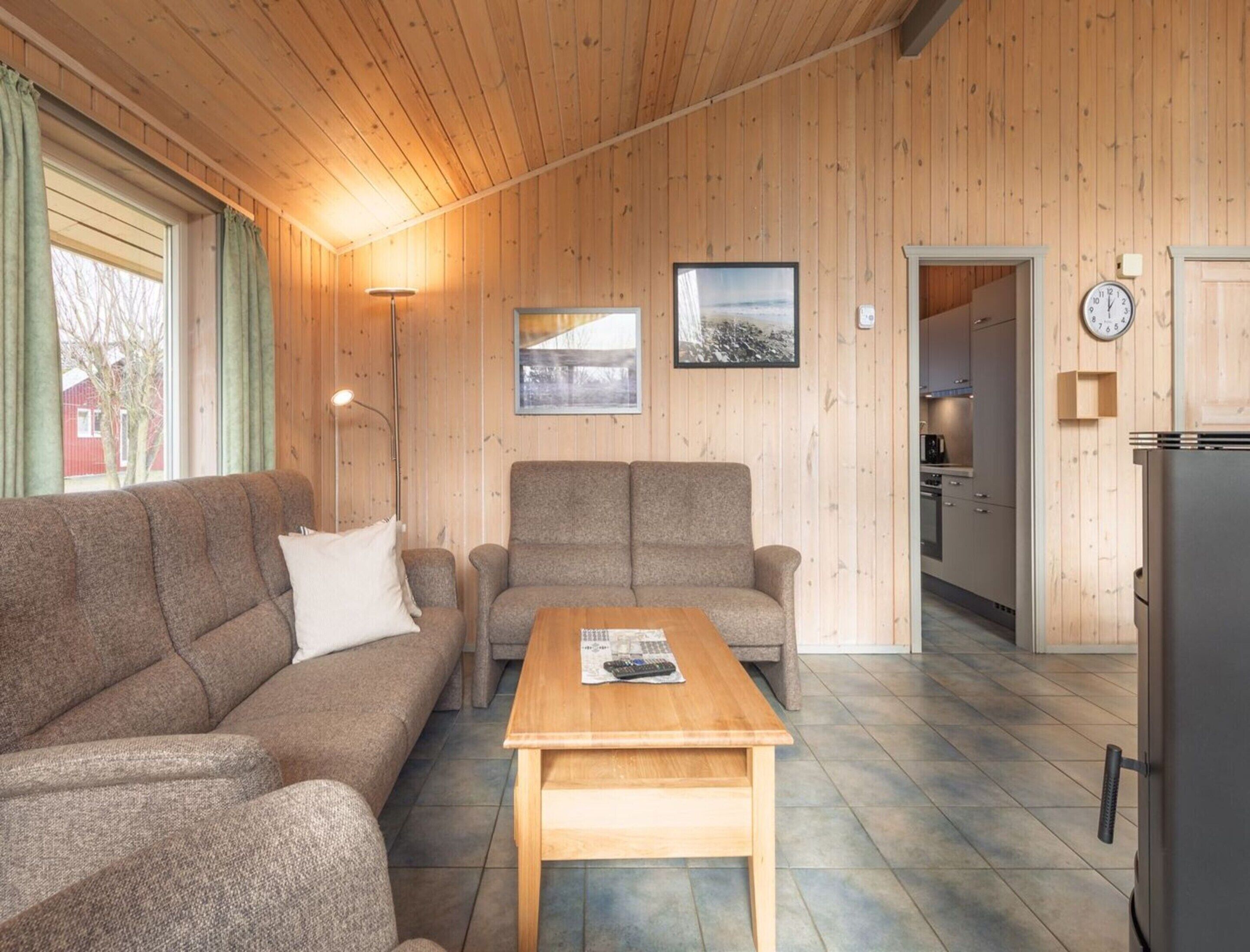 Un salon chaleureux avec un canapé, un fauteuil et une table basse, des murs en bois, une cuisine à l’arrière-plan et une horloge au mur.
