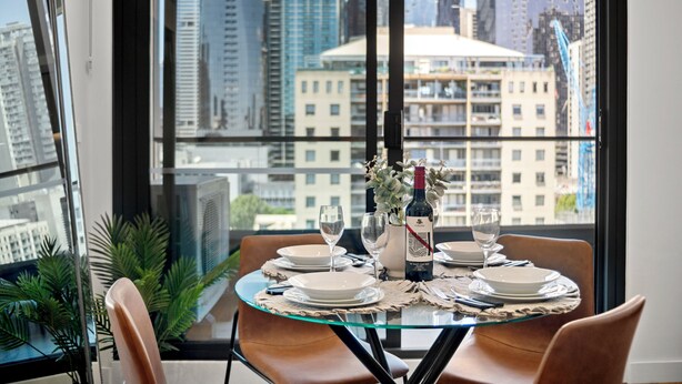Mesa de comedor para cuatro personas, con platos, vasos y una botella, ubicada en una habitación con un ventanal grande con vistas a la ciudad.