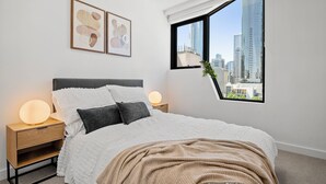 Un dormitorio con una cama, lámparas de noche, una mesita de noche, un ventanal con vistas a la ciudad y dos cuadros enmarcados en la pared.