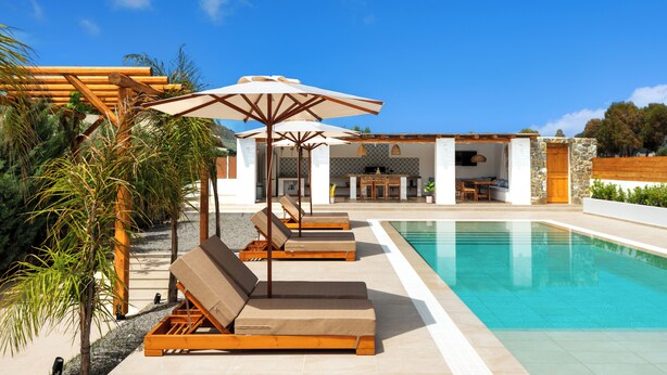 Un espace piscine avec des chaises longues et des parasols, un bâtiment en pierre doté d’une terrasse carrelée et un ciel d’un bleu limpide.