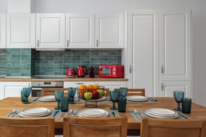 A modern kitchen with white cabinets, a wooden dining table set with plates and glasses, and a fruit bowl on the counter. - Johannes Luxury Villas (Rhodes)