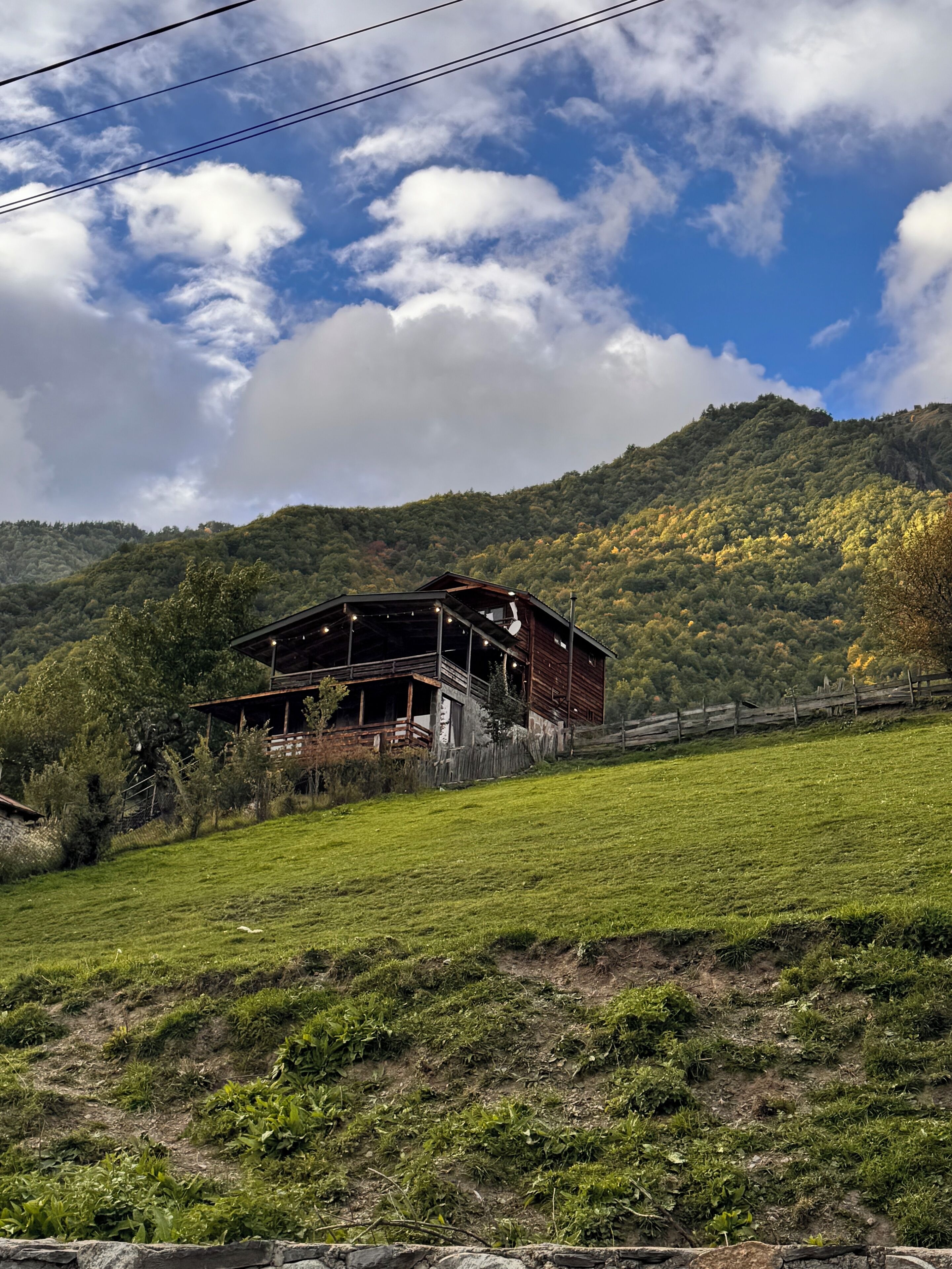 A rustic wooden cabin with a balcony, surrounded by a lush green field and a mountainous backdrop.