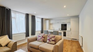 Un salon moderne avec un canapé et une table basse, une kitchenette dotée de meubles blancs et une fenêtre avec des rideaux.