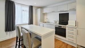 Une cuisine compacte dotée d’un îlot central, de meubles blancs, d’un four encastré et d’une fenêtre avec des rideaux.