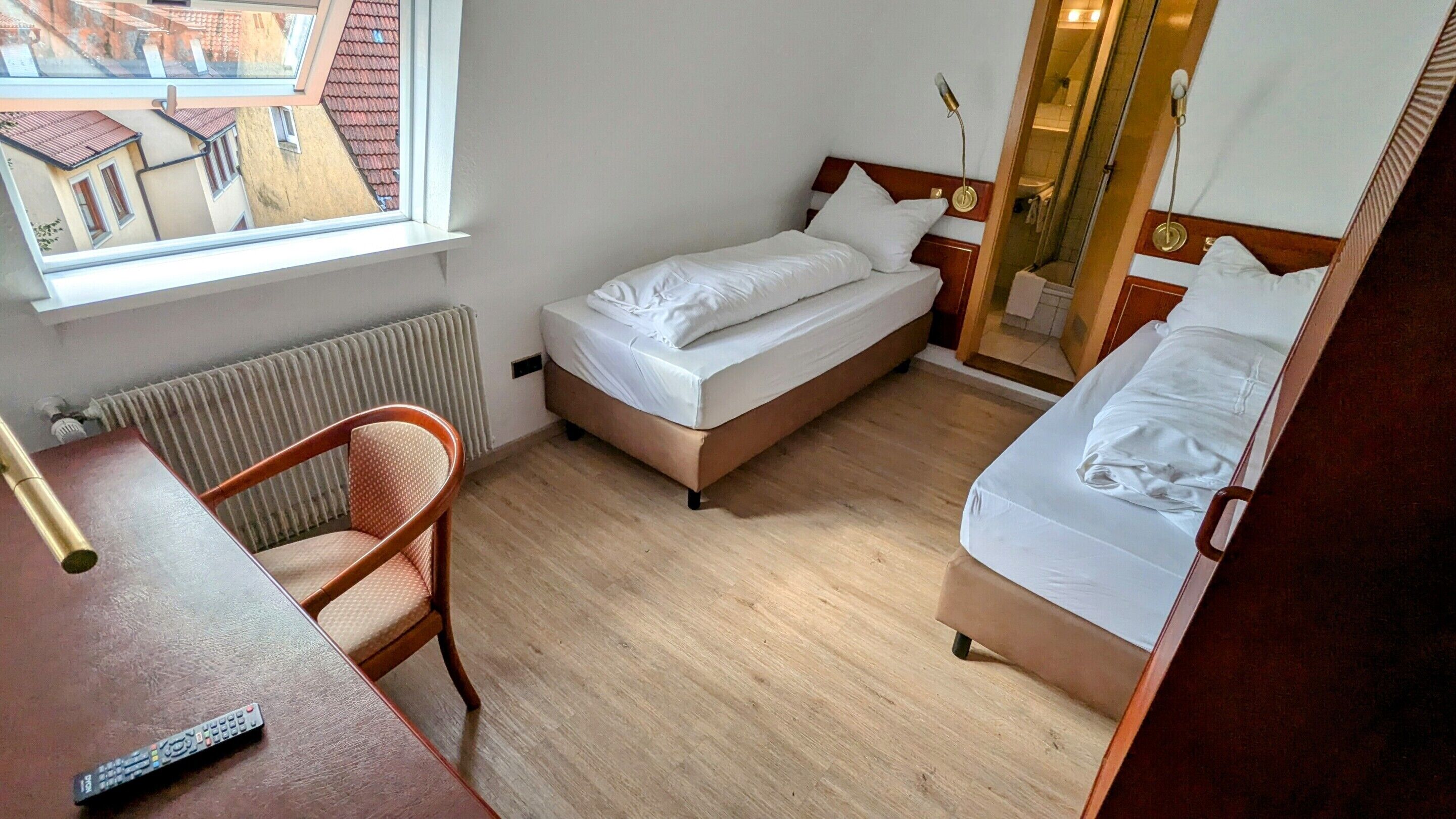A compact hotel room with a single bed, a chair, a desk, and a window overlooking a building.