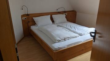 Una cama individual con estructura de madera y ropa de cama blanca en una habitación con suelo de madera y paredes blancas.