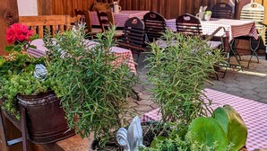 Un café al aire libre con mesas y sillas de madera, plantas en macetas y una pared de madera rústica.