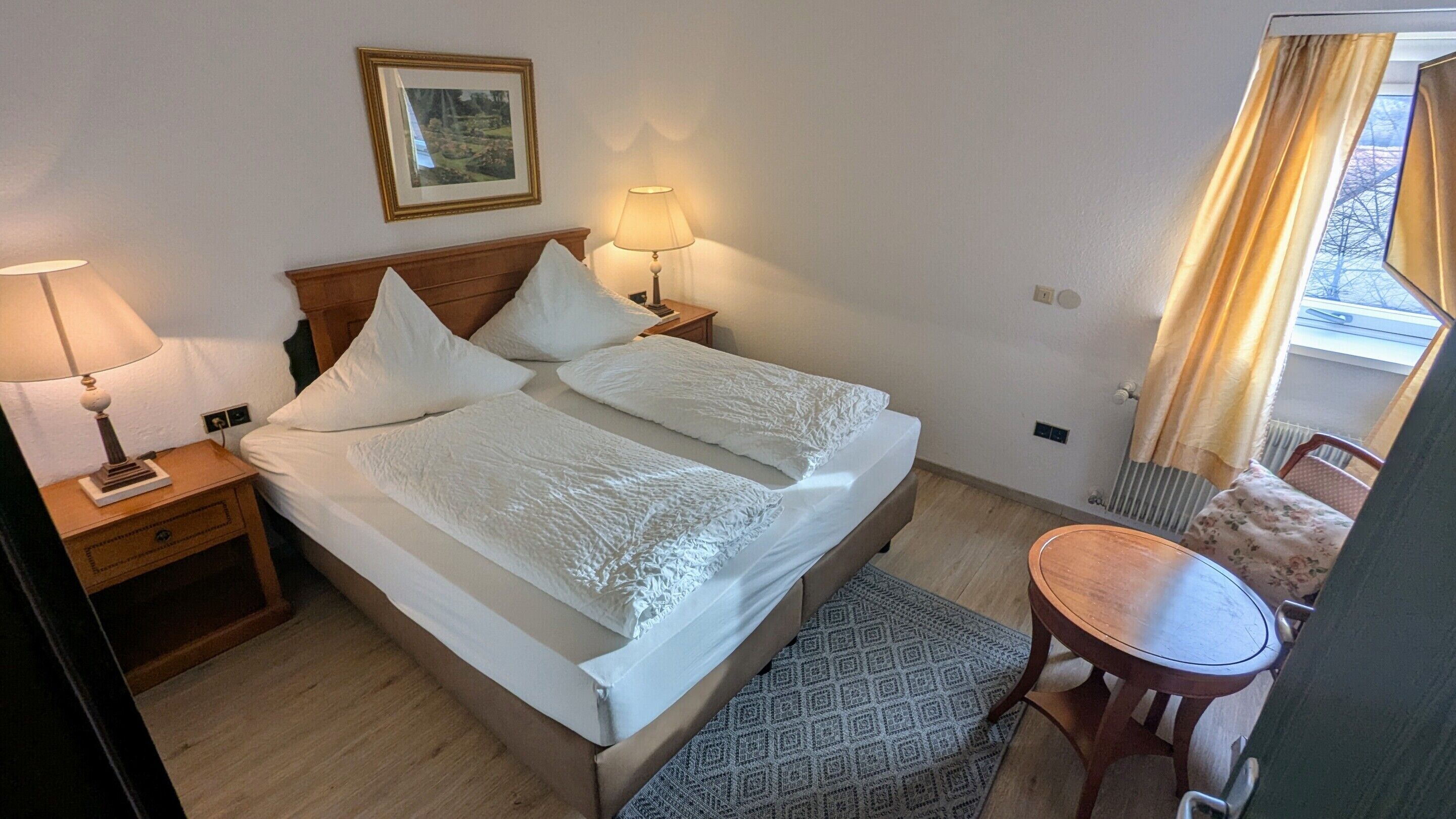 A double bed with white linens, two bedside lamps, a framed picture on the wall, a small wooden table, and a patterned rug.
