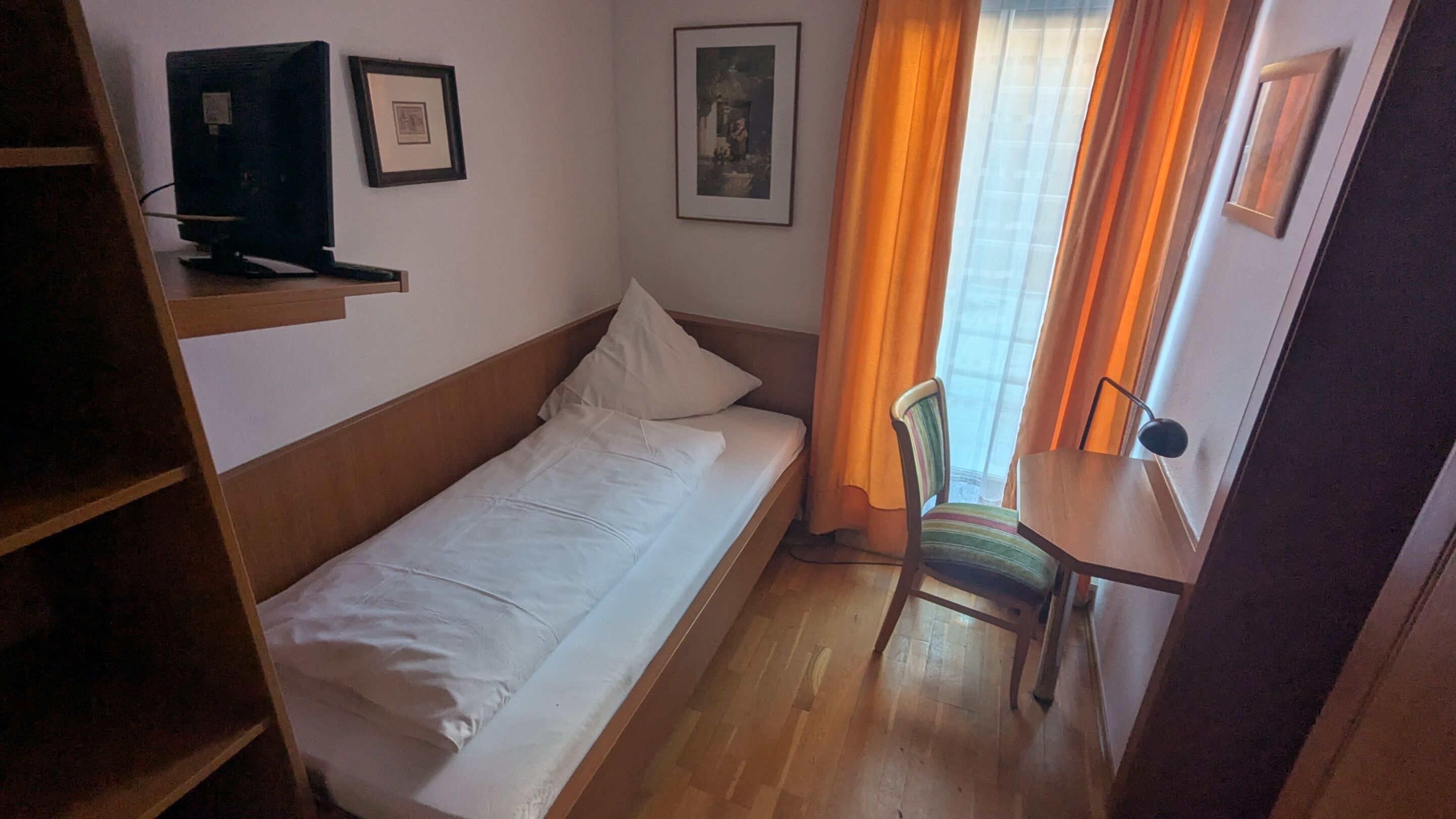 A small, single-bed room with a wooden headboard, a desk with a chair, and two framed pictures on the wall.