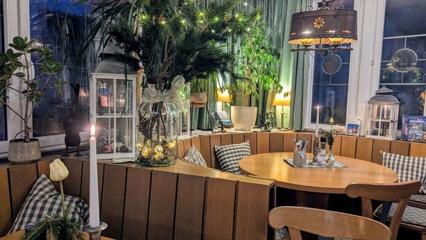 Un área de estar interior acogedora y bien iluminada, con paneles de madera, una mesa redonda y diversos elementos decorativos, como plantas y velas.