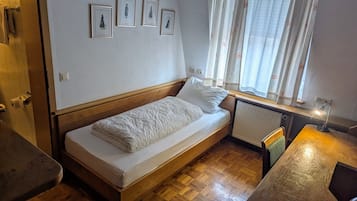 A small, single-bed room with a wooden headboard, a radiator, a window with curtains, and a small table with a lamp.
