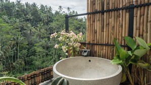Une grande fontaine ronde, ornée de motifs, située dans un jardin, avec une clôture en bambou et une végétation luxuriante en arrière-plan.