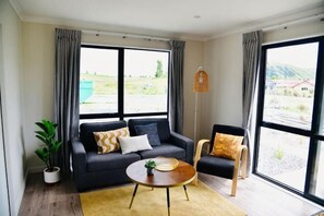 Una habitación con sofá, sillón, mesa de centro, lámpara de pie y ventanas grandes con cortinas.