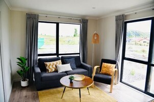 Una habitación con sofá, sillón, mesa de centro, lámpara de pie y ventanas grandes con cortinas.