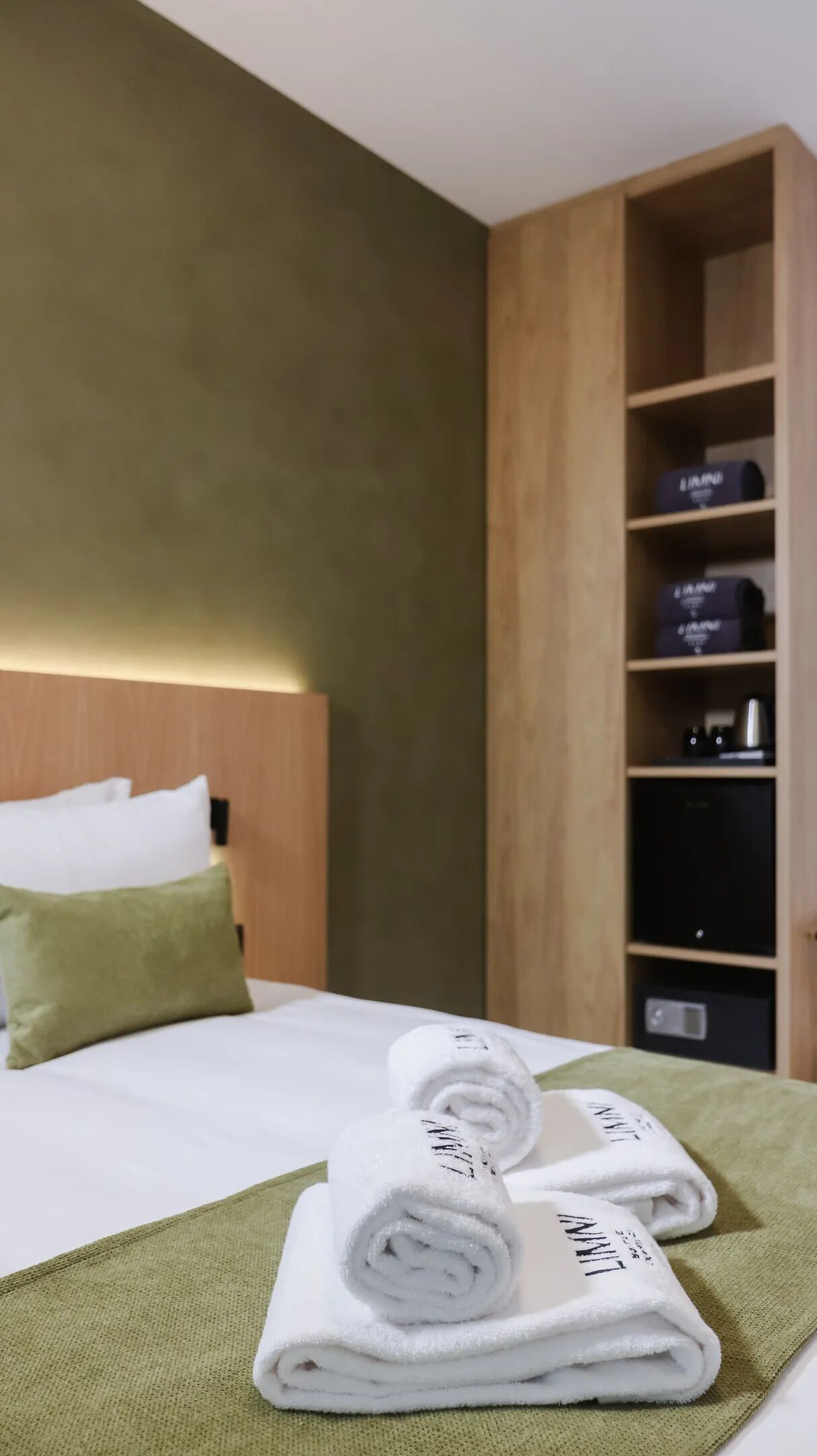 A hotel room with a bed, green towels, and a wooden wardrobe with storage boxes.