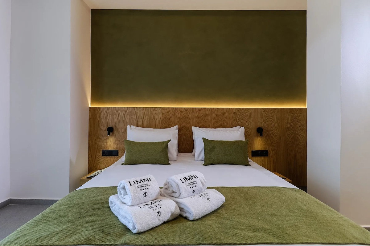 A hotel room with a wooden headboard, two white pillows, a green throw blanket, and two folded white towels with 'LIMNI' branding.