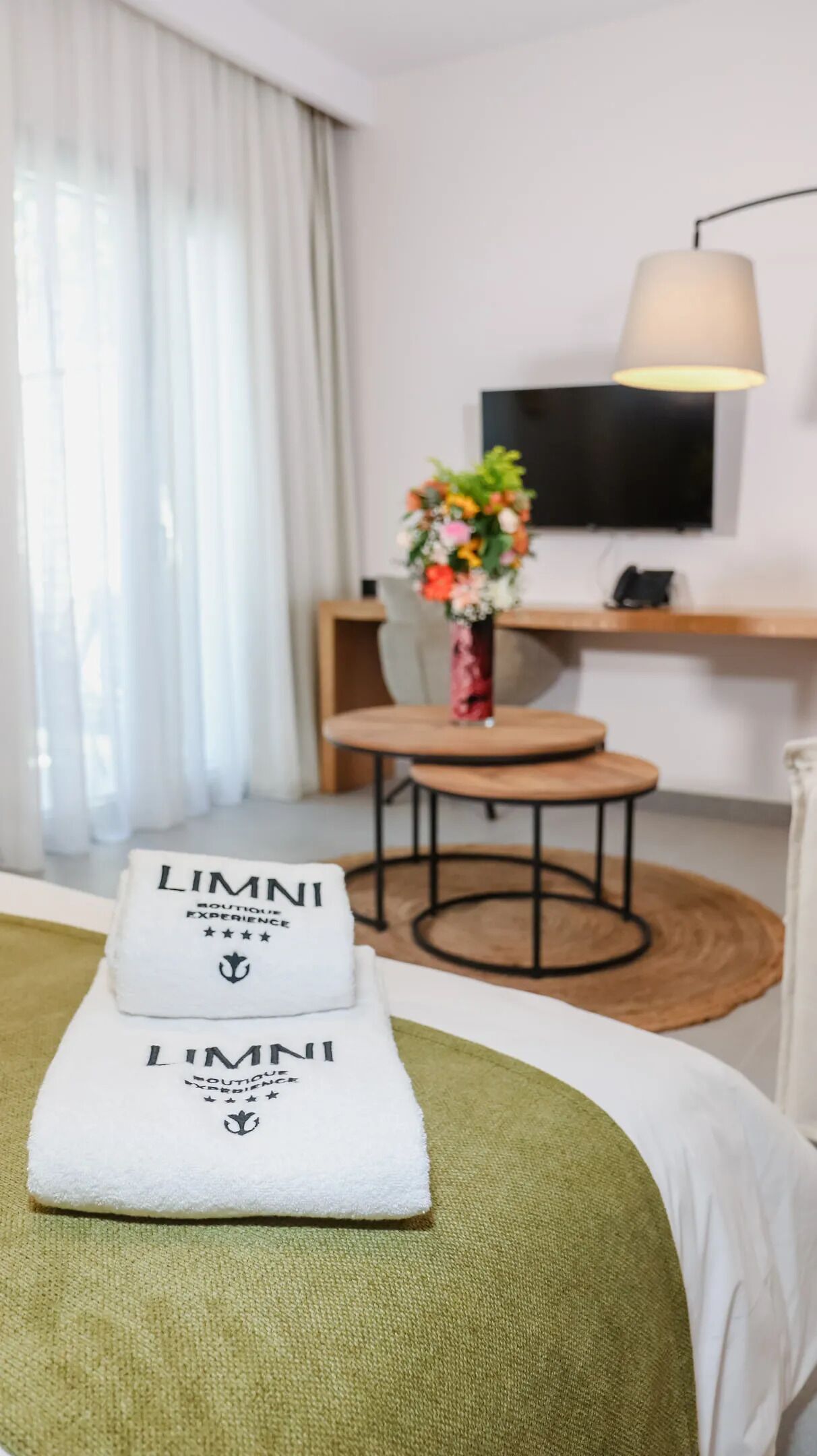 A hotel room with a bed, a green throw, a white pillow with 'LIMNI' branding, a small table with a vase of flowers, a chair, and a TV mounted on the wall.