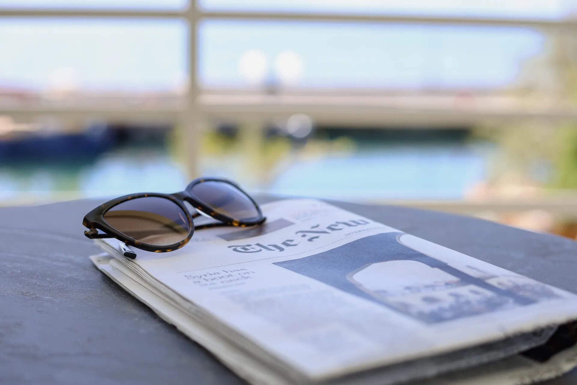 Une paire de lunettes posée sur un journal, avec en arrière-plan flou une étendue d’eau et un pont.